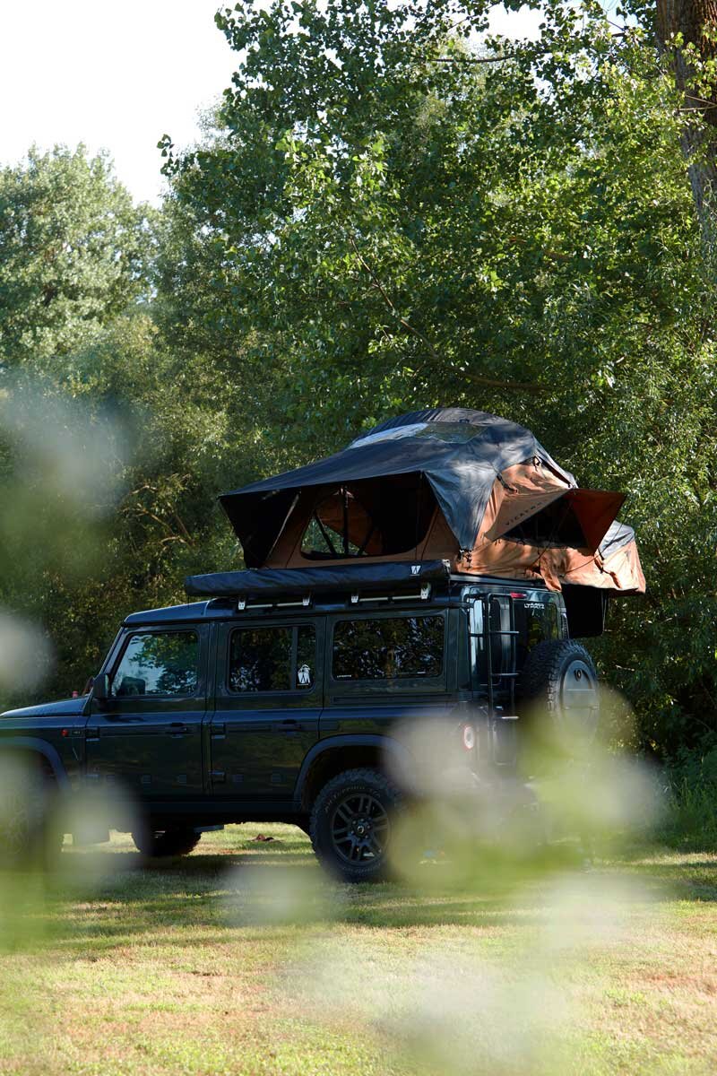 Two girls lay together in their rooftop tent, their dog looks up at them. Around the scene, there is camping-gear like chairs and a table.